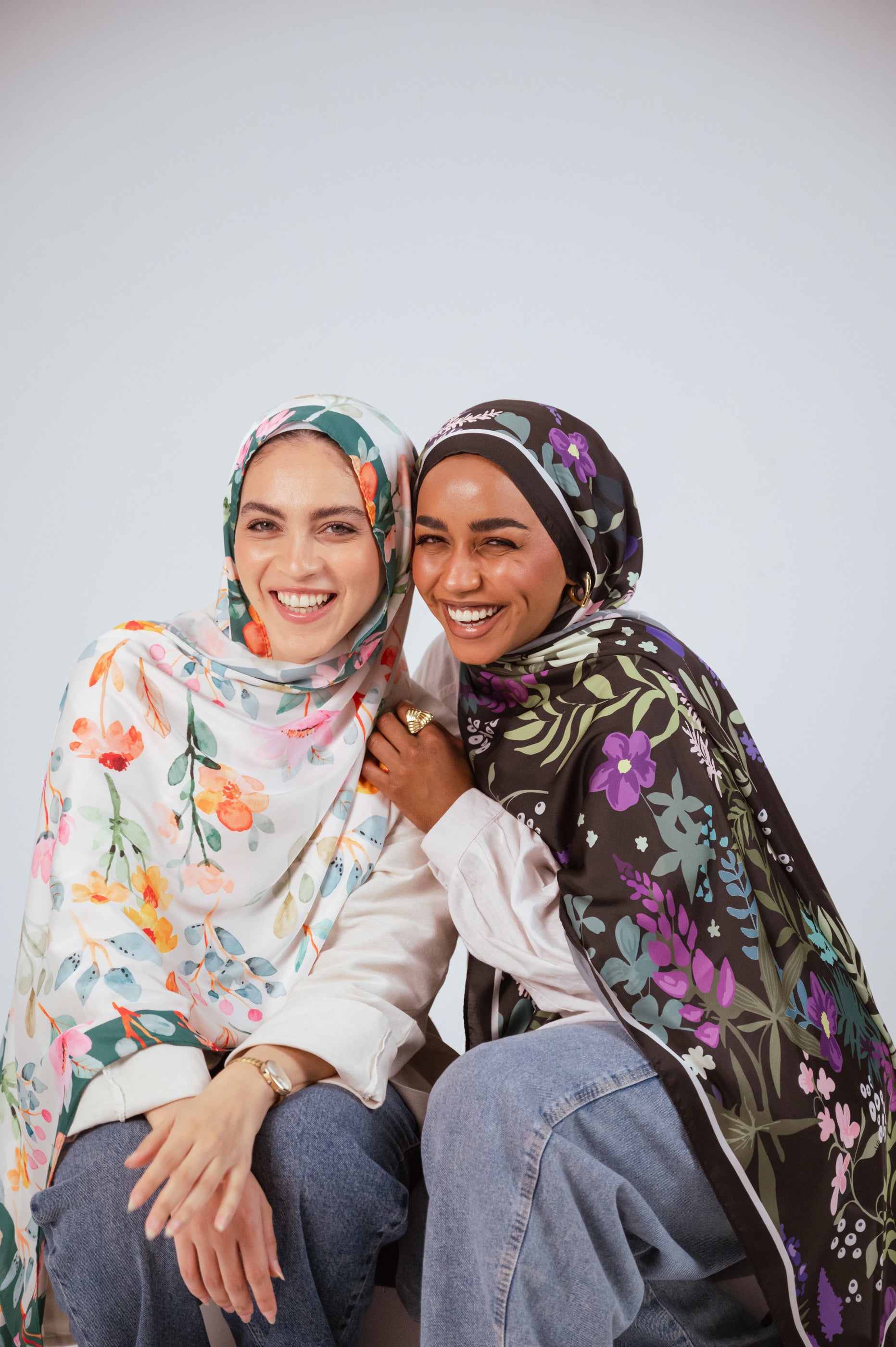 Models wearing the Andalus Drape and Blooms of Hope satin hijabs, smiling warmly while sitting close together.