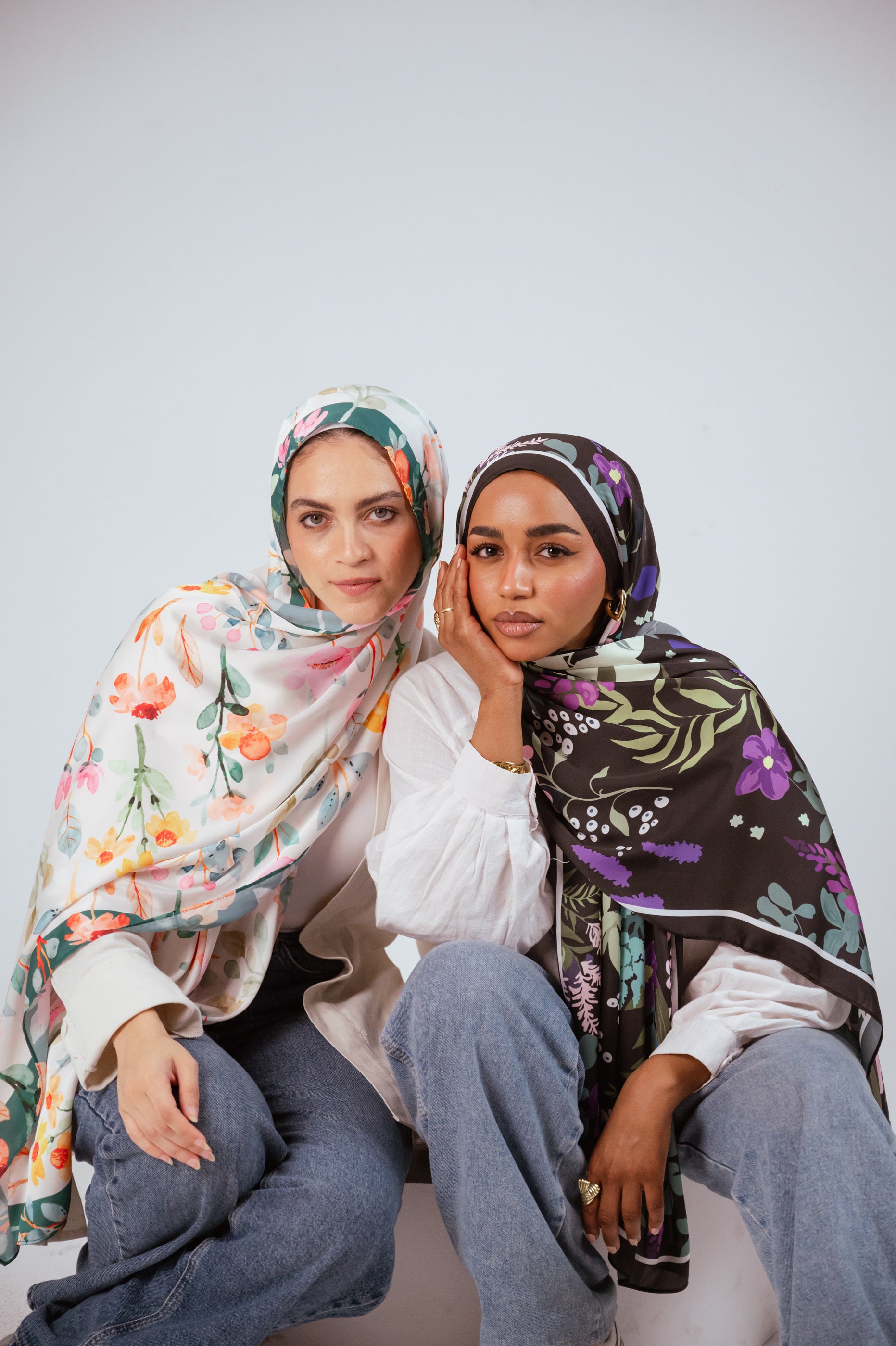 Models seated wearing Garden of Whispers and Blooms of Hope satin hijabs, showcasing print contrast and drape.