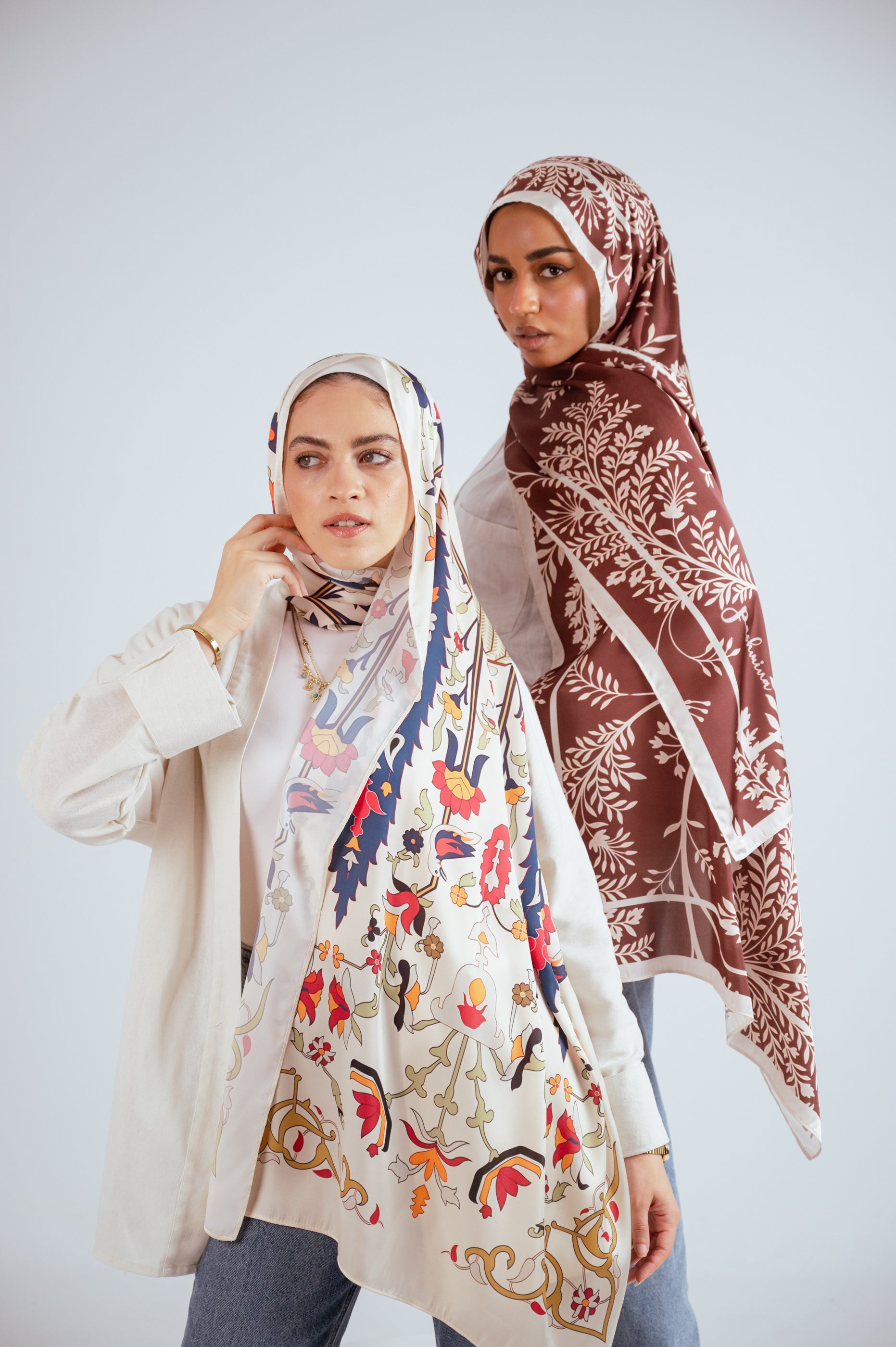 Close-up shot of two women wearing Andalus Drape Satin and Rose of Petra Satin hijabs, showcasing the detailed artwork.