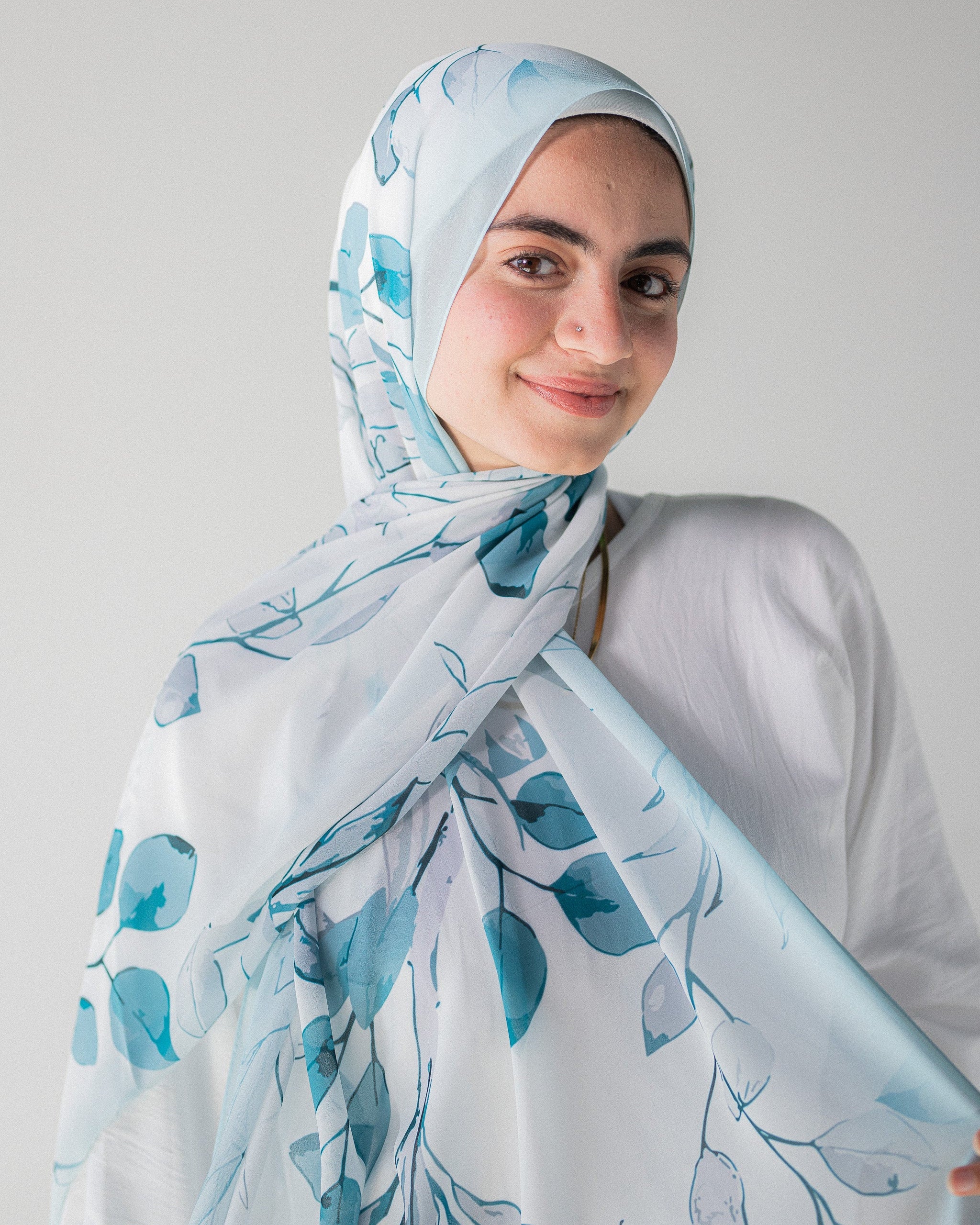 Modest Muslim young woman wearing a light blue hijab with floral patterns on a plain background
