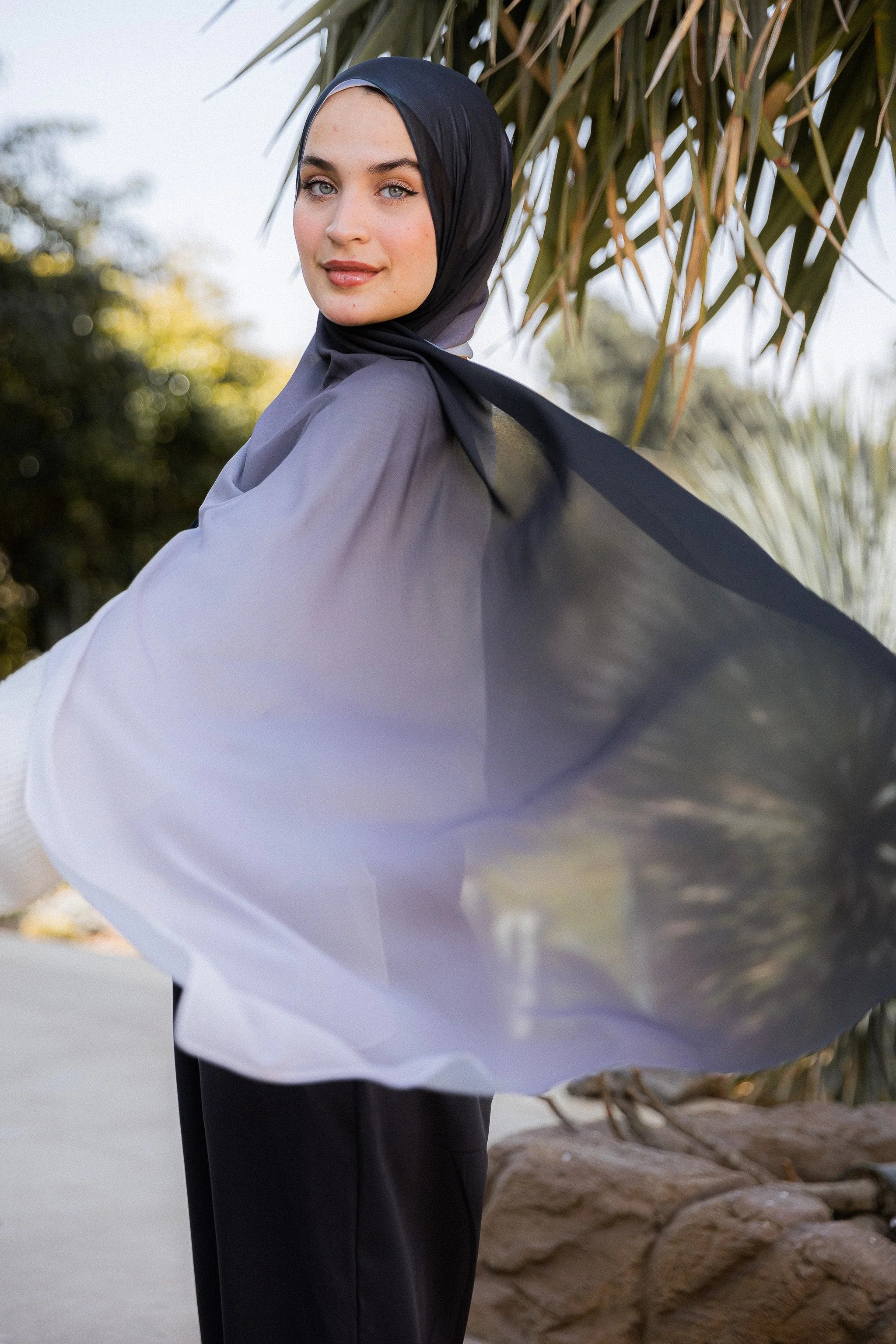 Lunar Mist chiffon hijab in motion, revealing the full black-to-silver ombré gradient against a palm backdrop