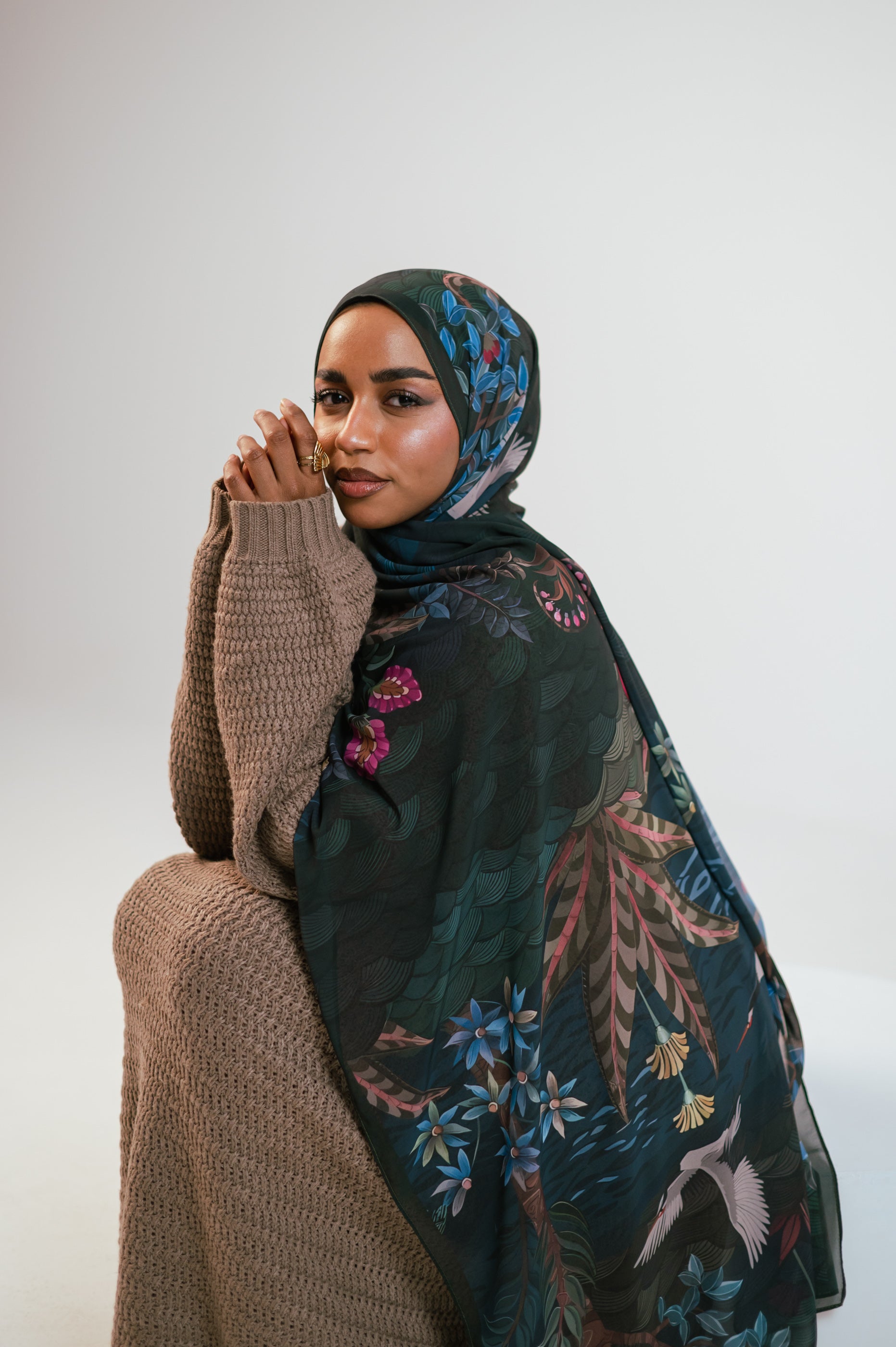 Model seated with hands together wearing Secret Garden Luxury Chiffon Hijab in deep green floral print.