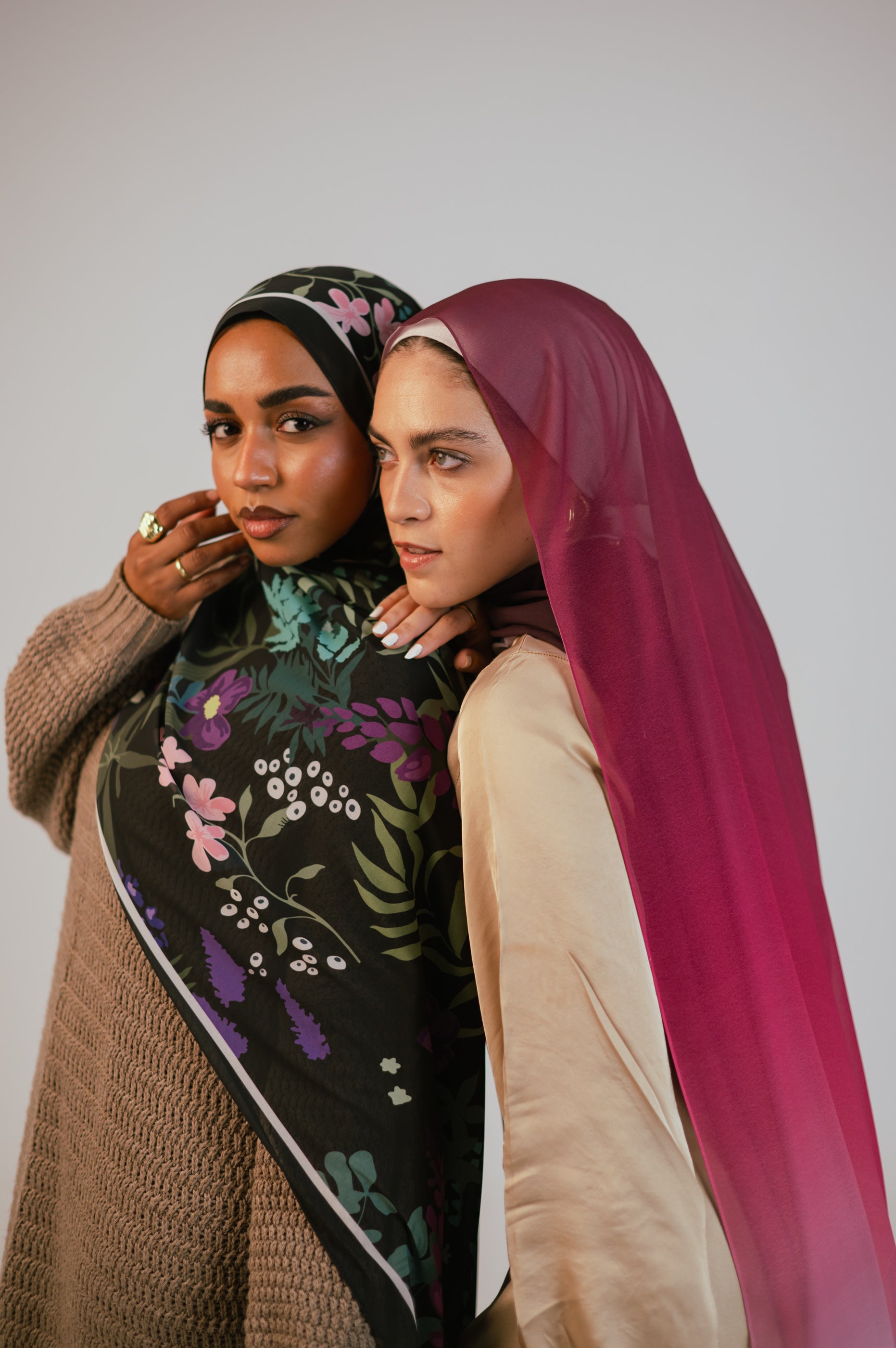 Two women wearing JAIDA luxury chiffon hijabs — Garden of Whispers in black floral and Wisteria Sunset in pink ombré — styled together in an elegant duo pose.