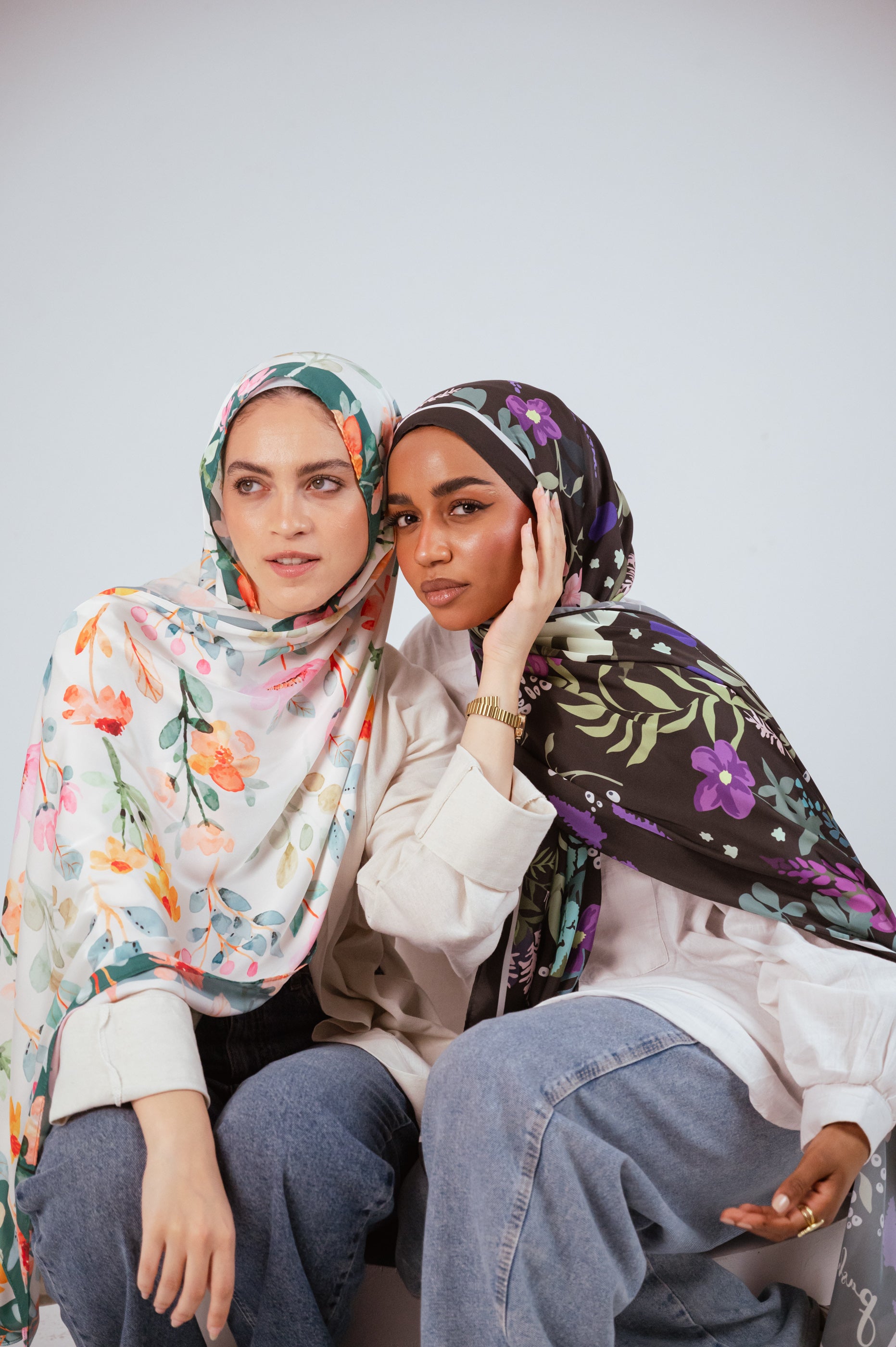 Two models wearing the Andalus Drape and Blooms of Hope satin hijabs, posing side by side in a soft, elegant seated composition.