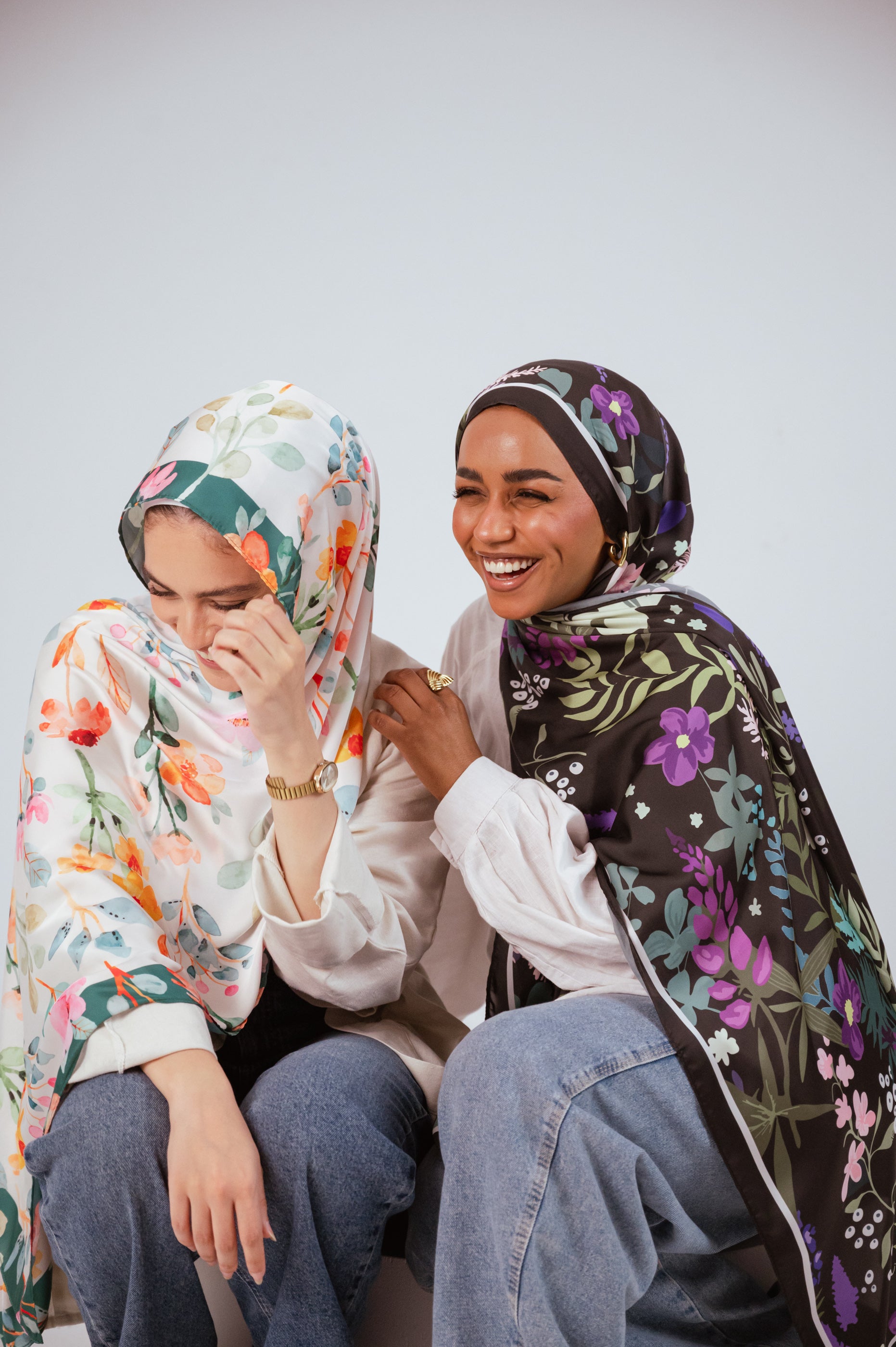 Models laughing together while wearing the Andalus Drape satin hijab and the Blooms of Hope satin hijab in a relaxed seated pose.