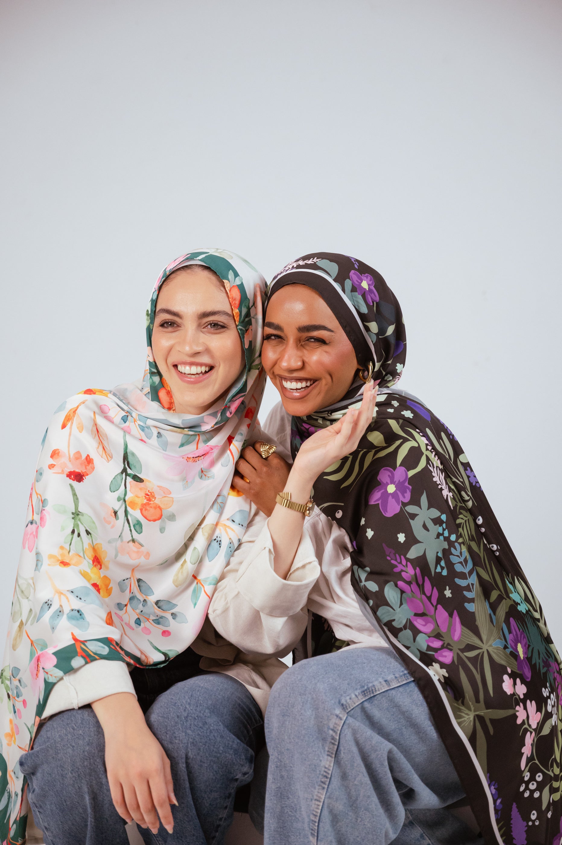 Two models wearing the Andalus Drape satin hijab and Blooms of Hope satin hijab, sitting together and laughing in a joyful, bright studio setting.