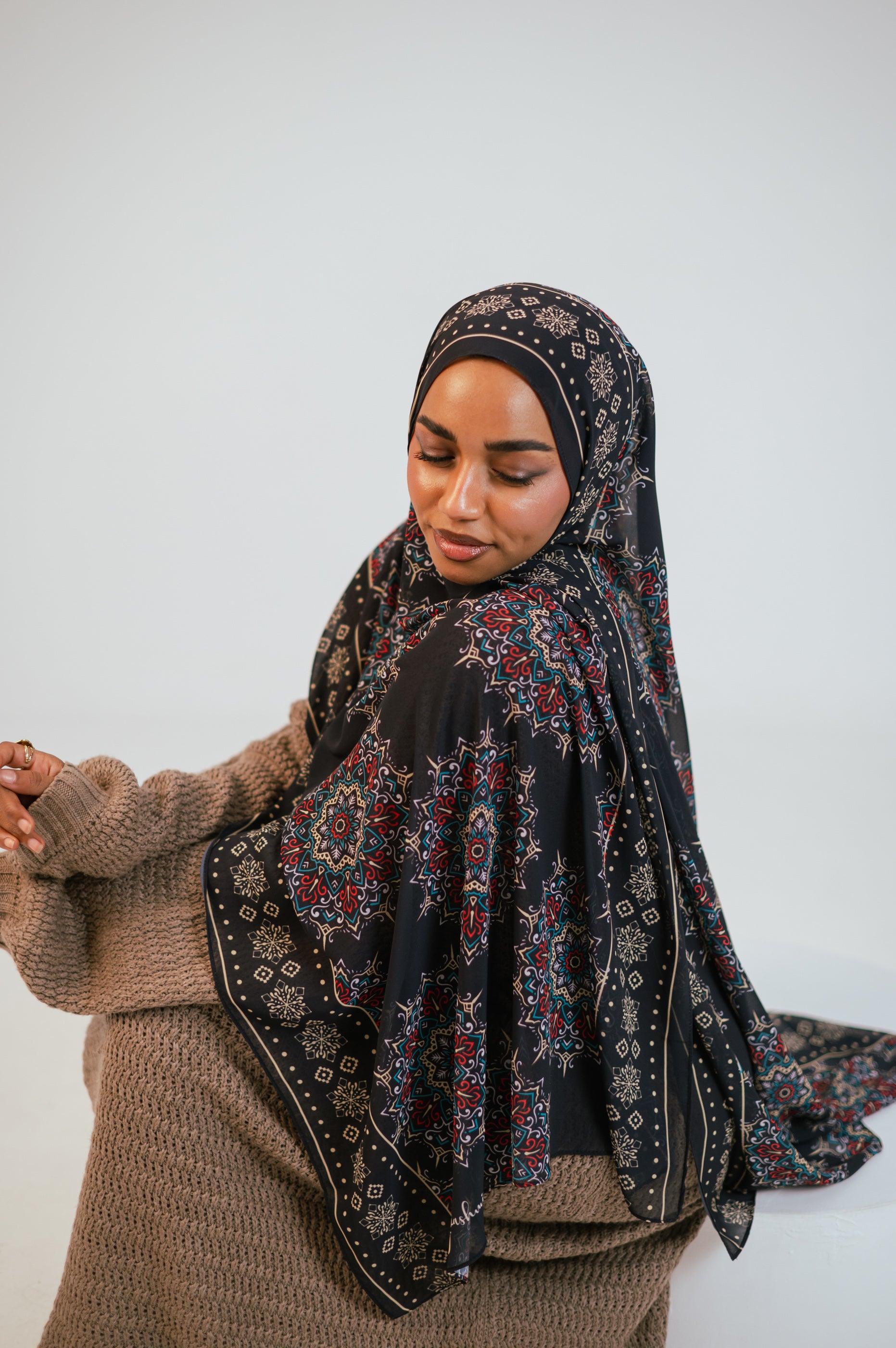 Seated editorial portrait featuring the Andalusia Luxury Chiffon Hijab styled with a textured knit dress, showcasing contrast and print clarity.
