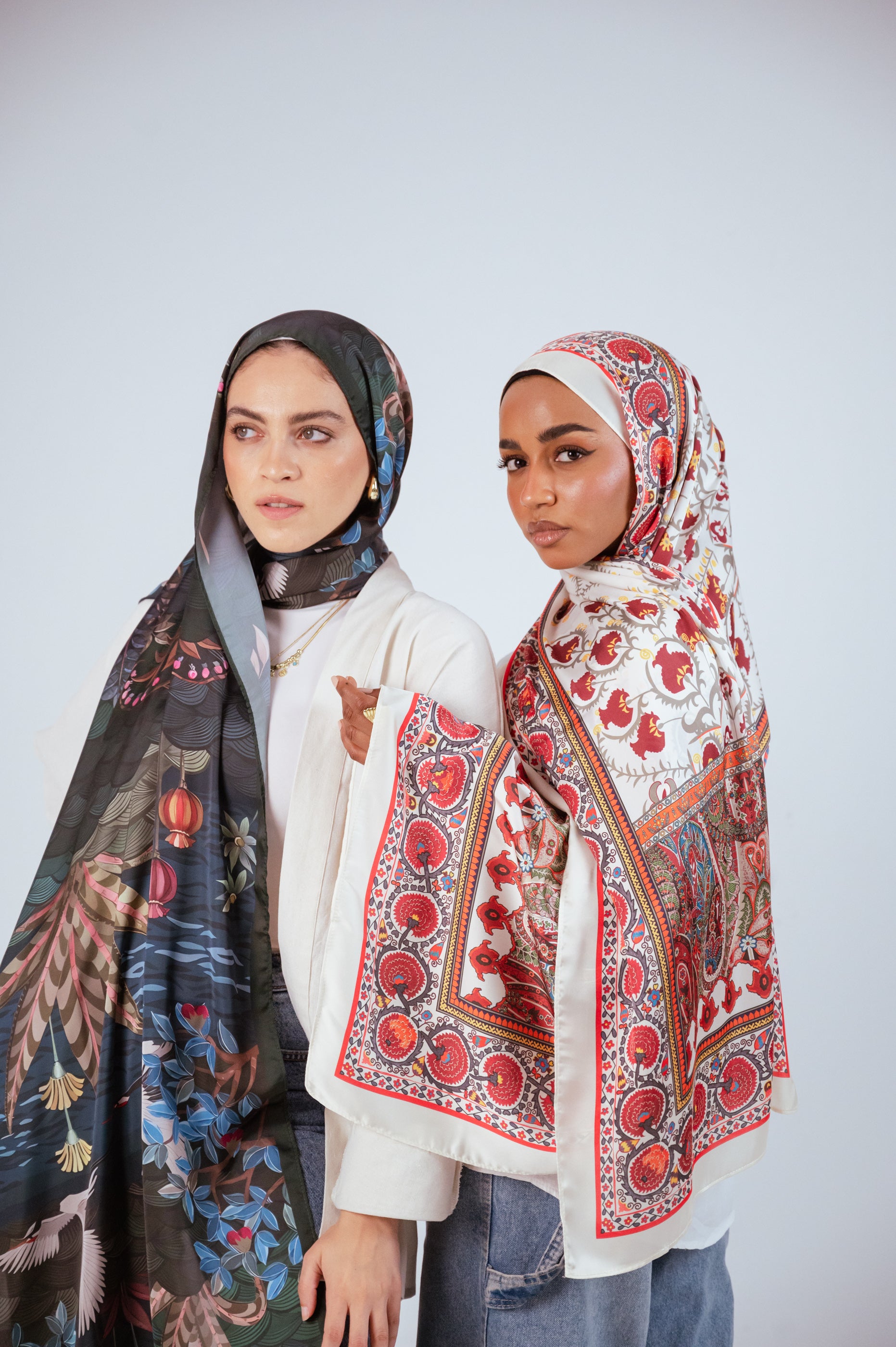 Two models in JAIDA hijabs posing together, contrasting the rich red motifs of Cairo Nights Satin with the moody botanical tones of Secret Garden Chiffon.