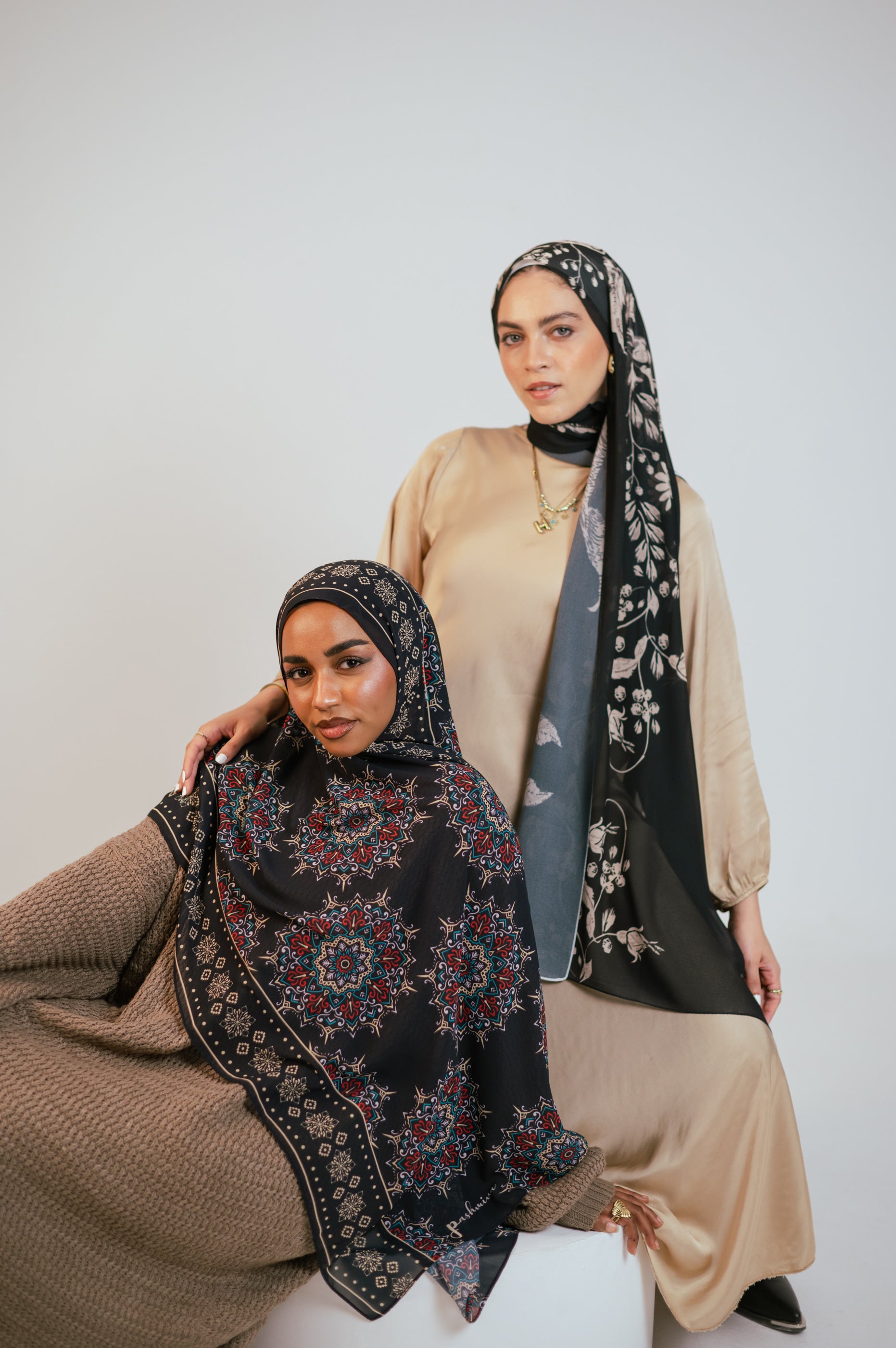 Two models wearing Petal Shadows luxury chiffon hijab and a patterned chiffon hijab, seated in a studio setting showing elegant drape and floral detailing.