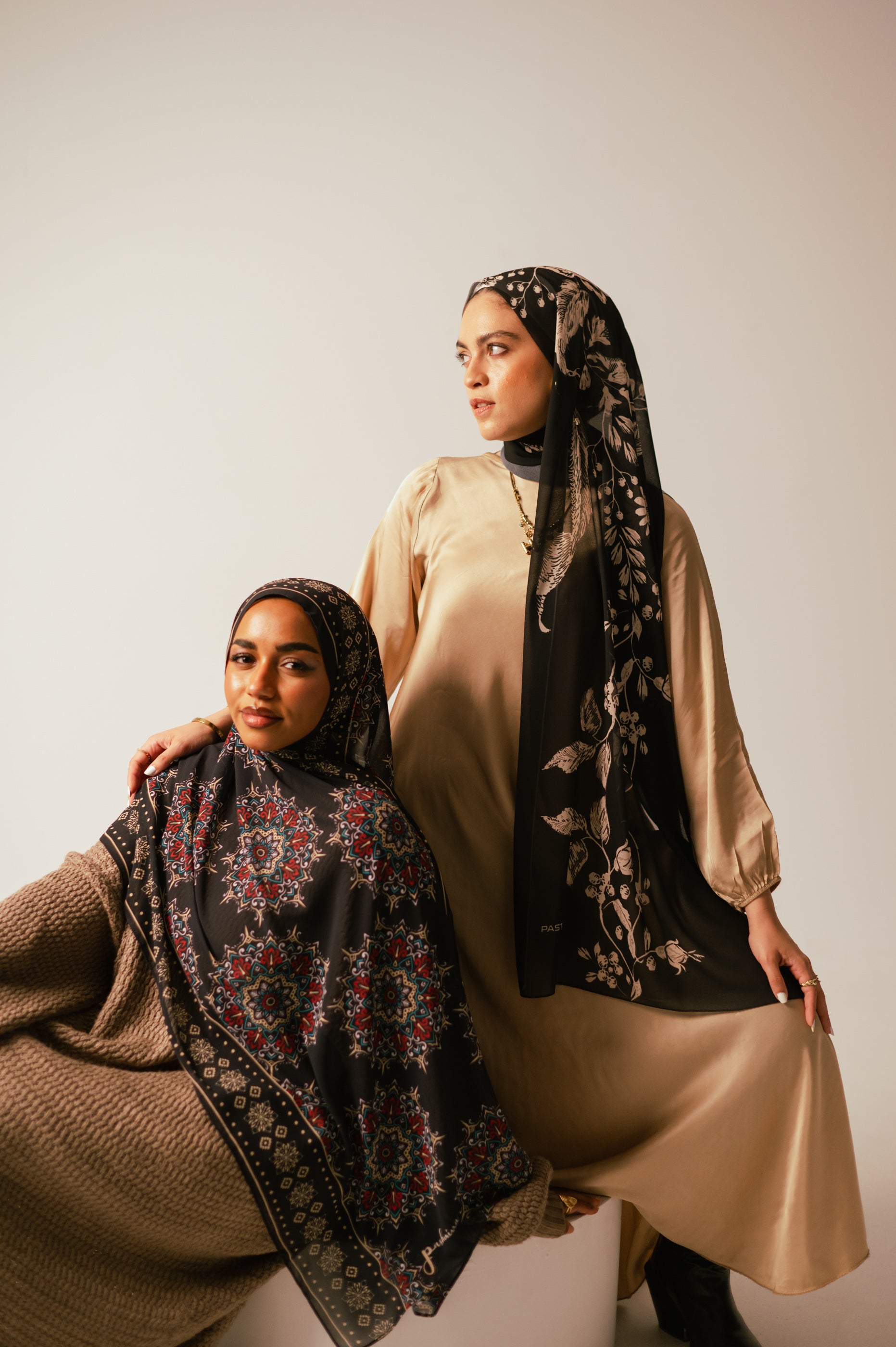 Two models in warm studio lighting wearing Petal Shadows luxury chiffon hijab alongside a geometric-print chiffon hijab, showcasing full drape and modest styling.